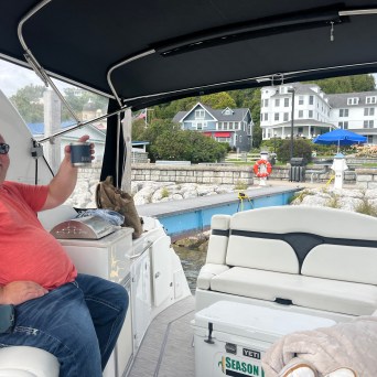Man in sunglasses on boat holding a drink, with waterfront houses in the background.