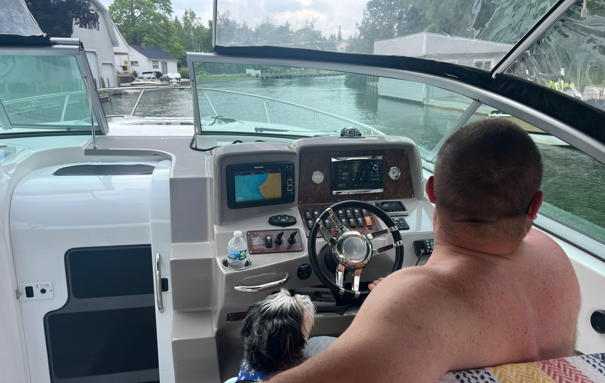 A man and a small dog sit at a boat's steering area, facing a lake view.