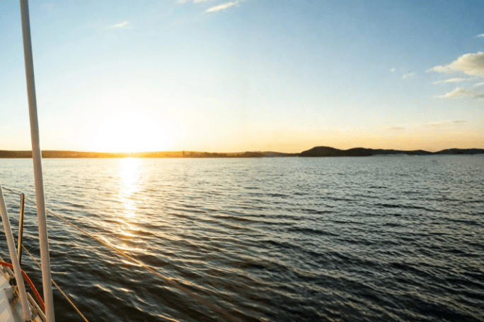Sailboat at sunset on calm water with distant hills.